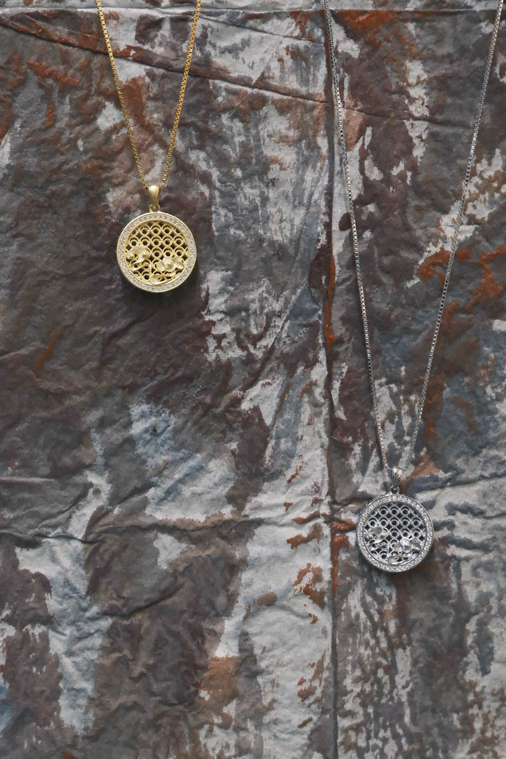 Two gold and silver necklaces with intricate pendants on a textured surface.
